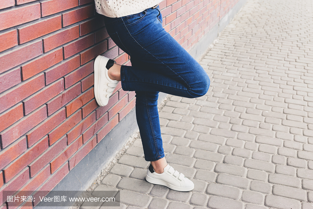 女孩站在蓝色牛仔裤和白色运动鞋旁边的墙上