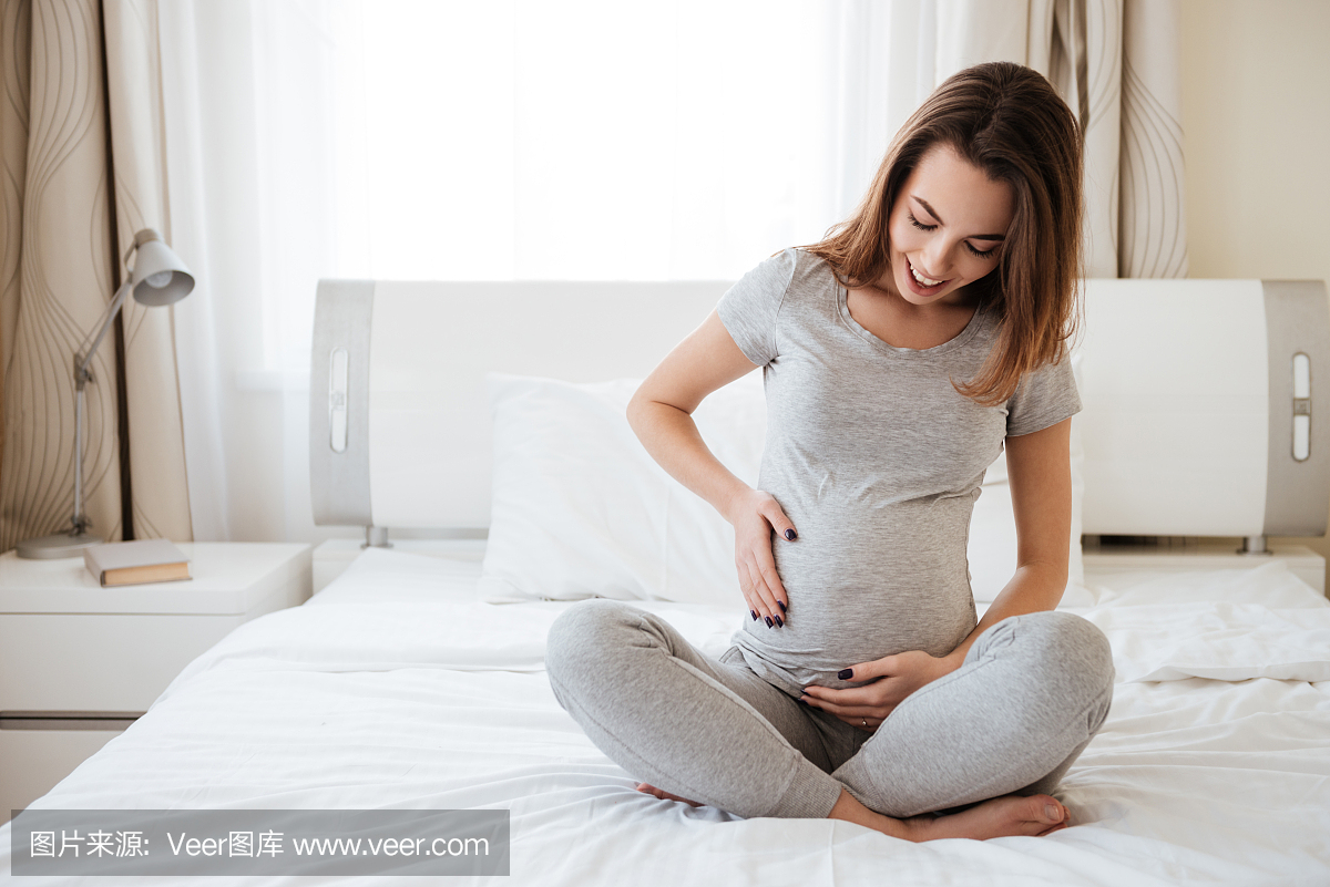 快乐的孕妇坐在床上,摸她的肚子