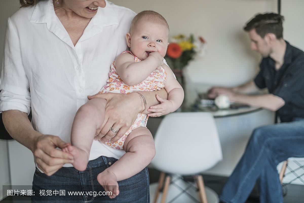 母亲抱着宝宝(2-5个月)和父亲在背景下工作