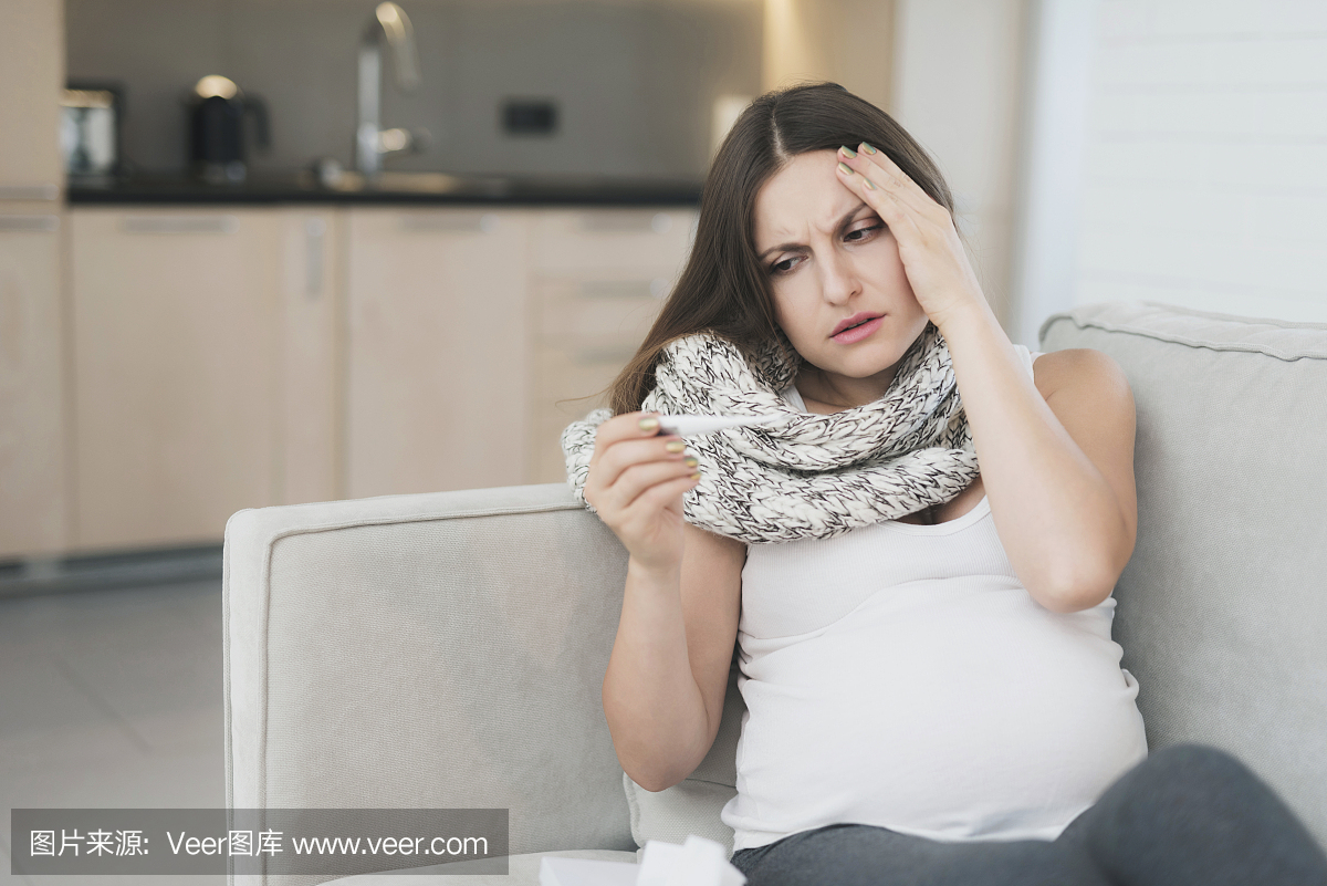 生病的孕妇坐在家里在沙发上。它用电子温度计