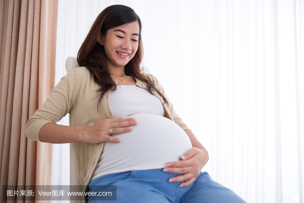 快乐怀孕的亚裔女人在坐着时触摸她的大肚子